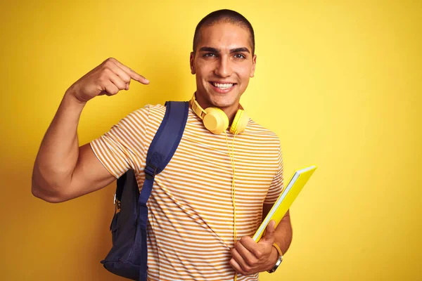 若いです学生男身に着けていますヘッドフォンとバックパック上の黄色隔離された背景とともに驚き顔指摘指へ彼自身 — ストック写真