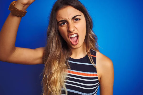 Young beautiful woman wearing stripes t-shirt over blue isolated background annoyed and frustrated shouting with anger, crazy and yelling with raised hand, anger concept