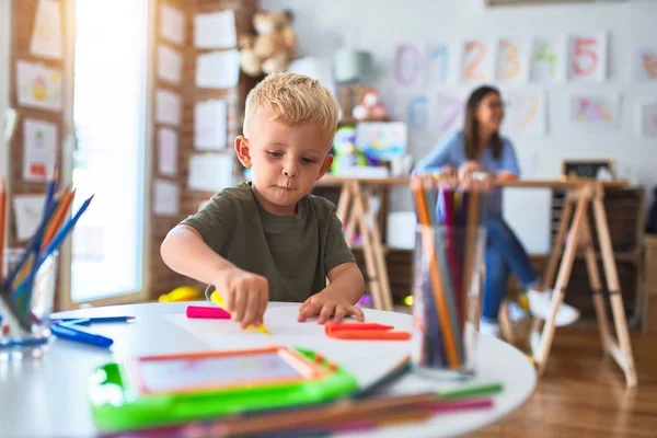 年轻的高加索孩子和老师在学校玩耍 游戏室的母亲和儿子用彩色铅笔画了一幅画 背景上的年轻女子坐在桌子上 — 图库照片