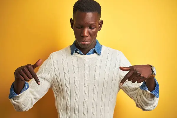 アフリカ系アメリカ人の男性はデニムシャツと白いセーターを身に着けています孤立した黄色の背景の上に下を向いて悲しいと動揺 指で方向を示す 不幸なと落ち込んで — ストック写真