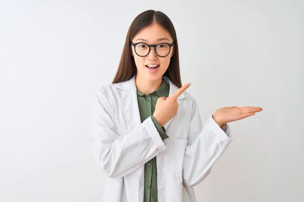 Young chinese scientist woman wearing coat and glasses over isolated white background amazed and smiling to the camera while presenting with hand and pointing with finger.