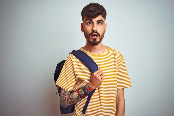 Young student man with tattoo wearing backpack standing over isolated white background scared in shock with a surprise face, afraid and excited with fear expression