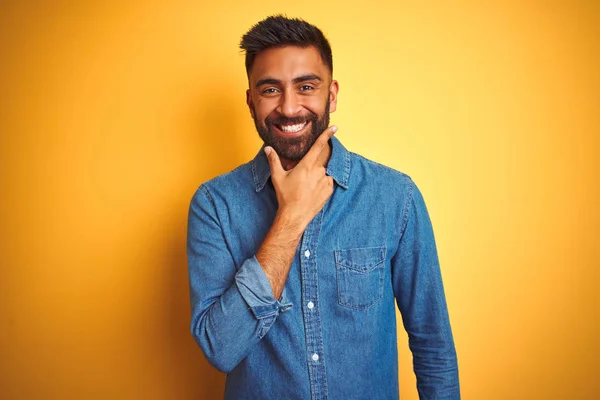 独立した黄色の背景の上にデニムシャツを着た若いインド人男性は カメラが交差した腕と手であごを上げて笑っていることに自信を持っている 肯定的な考え — ストック写真