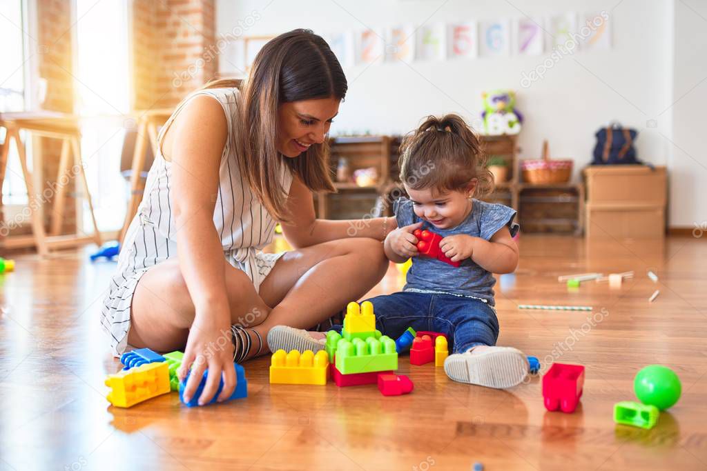 Young beautiful teacher and toddler playing with building blocks toy at kindergarten