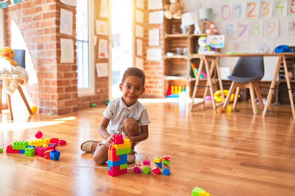 Beautiful african american toddler playing with wooden blocks train toy around lots of toys at kindergarten