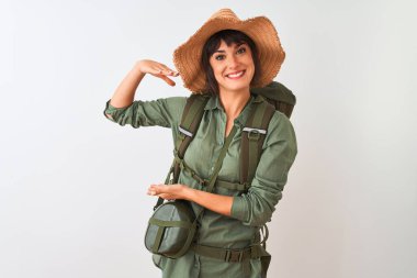 Sırt çantası şapkası ve su matarası takan yürüyüşçü kadın izole edilmiş beyaz arka plan üzerinde büyük ve büyük boyut işareti, ölçü sembolü gösteren el hareketleriyle. Kameraya bakıp gülümsüyor. Ölçüm konsepti.