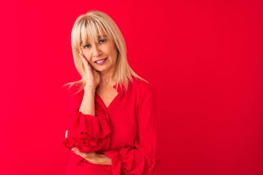 Soyutlanmış kırmızı arka plan üzerinde duran zarif gömlekli orta yaşlı bir kadın kollarını kavuşturarak depresyondan sıkılmış ve yorgun göründüğünü düşünüyor..