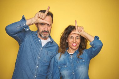 Güzel orta yaşlı bir çift, izole edilmiş sarı arka planda dikiliyorlar. Alnında parmakları olan insanlarla dalga geçiyorlar. Ezik jestler yapıp alay ediyorlar..