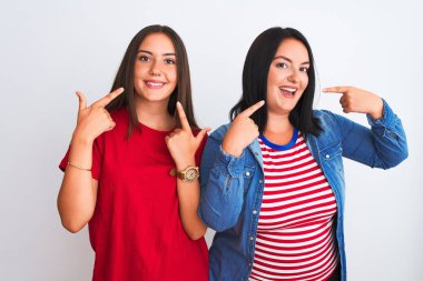 İzole edilmiş beyaz arka plan üzerinde sıradan kıyafetler giyen genç ve güzel kadınlar neşeli bir şekilde gülümseyip parmaklarını ve ağzını gösteriyorlar. Diş sağlığı konsepti.