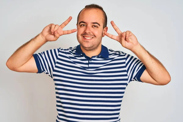Sıradan çizgili polo giyen genç adam izole edilmiş beyaz arka planda duruyor. Yüzünde parmakları olan barış sembolü yapıyor. Neşeli bir şekilde zafere gülümsüyor.