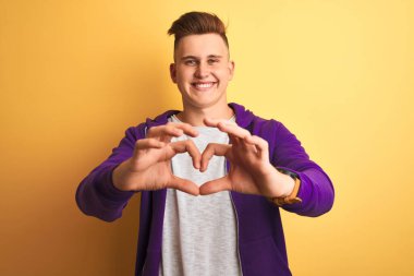 Mor kazak giyen genç yakışıklı adam izole edilmiş sarı arka planda duruyor. Aşkla gülümsüyor, elleriyle kalp sembolü yapıyor. Romantik konsept.