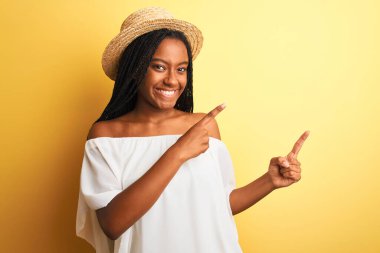 Beyaz tişörtlü ve şapkalı genç Afro-Amerikalı kadın izole edilmiş sarı arka planda gülümsüyor ve kameraya bakıyor..