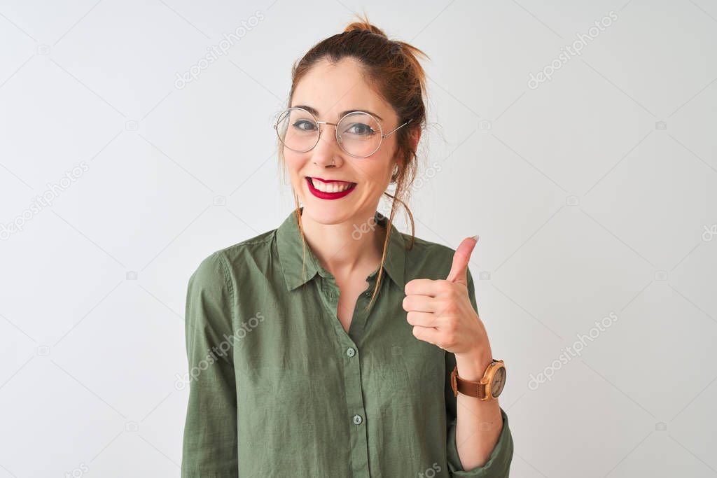 Redhead woman wearing green shirt and glasses standing over isolated white background doing happy thumbs up gesture with hand. Approving expression looking at the camera with showing success.