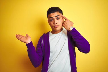 Mor eşofman üstü giyen genç Brezilyalı adam izole edilmiş sarı arka planda duruyor. Kafası karışmış ve avuç içi açık bir şekilde fotokopi alanını gösteriyor ve alnını işaret ediyor. Bunu bir düşün..