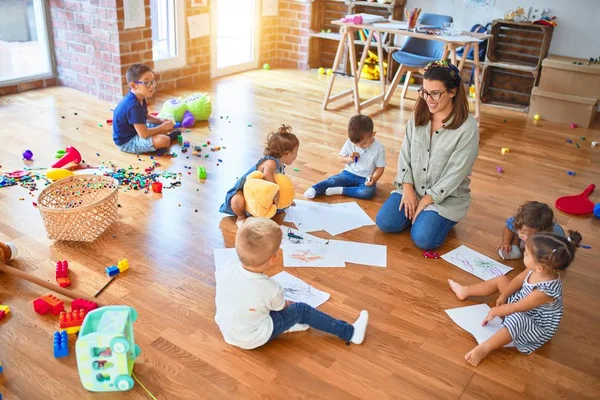 幼稚園のおもちゃの周りに紙や鉛筆を使って床に座っている子供たちの美しい先生とグループ — ストック写真