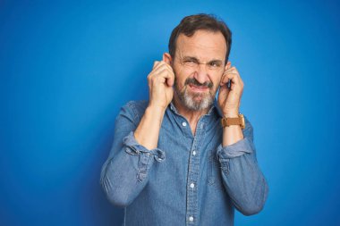Yakışıklı, orta yaşlı, gri saçlı, izole edilmiş mavi arka planı olan, yüksek sesli müzik sesi için parmaklarıyla kulaklarını kapatan yaşlı bir adam. Sağır kavramı.