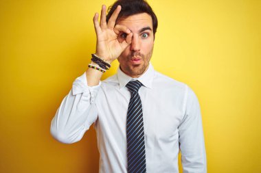 Zarif gömlek ve kravat takan genç yakışıklı iş adamı, izole edilmiş sarı arka plan üzerinde şaşırmış yüz ve parmaklarından bakan gözle şok edici hareketler yapıyor. İnançsız ifade.