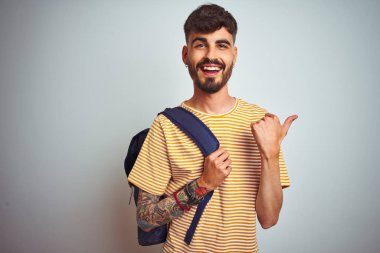 Sırt çantasında dövmesi olan genç bir öğrenci izole edilmiş beyaz arka plan üzerinde duruyor ve gülümseyen bir yüzle baş parmağıyla onu gösteriyor.