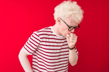 İzole edilmiş kırmızı arka plan üzerinde çizgili tişört ve gözlük takan genç sarışın adam kendini kötü hissediyor ve soğuk algınlığı veya bronşitin belirtisi olarak öksürüyor. Sağlık hizmetleri kavramı.