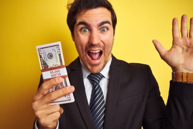 Takım elbiseli genç yakışıklı işadamı, izole edilmiş sarı arka planda dolar tutuyor. Çok mutlu ve heyecanlı, kazanan yüz ifadesi zaferi büyük bir gülümsemeyle kutluyor ve ellerini kaldırıyor.