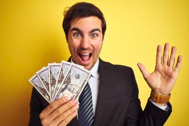 Takım elbiseli genç yakışıklı işadamı, izole edilmiş sarı arka planda dolar tutuyor. Çok mutlu ve heyecanlı, kazanan yüz ifadesi zaferi büyük bir gülümsemeyle kutluyor ve ellerini kaldırıyor.