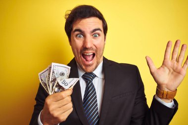 Takım elbiseli genç yakışıklı işadamı, izole edilmiş sarı arka planda dolar tutuyor. Çok mutlu ve heyecanlı, kazanan yüz ifadesi zaferi büyük bir gülümsemeyle kutluyor ve ellerini kaldırıyor.
