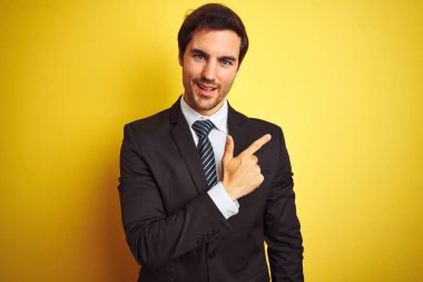 Genç yakışıklı iş adamı takım elbise ve kravat takıyor, sarı arka planda mutlu bir şekilde duruyor, yüzünde mutlu ve doğal bir ifadeyle elleri ve parmaklarını gösteren bir gülümseme var.