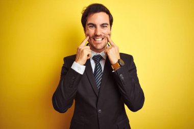 Genç yakışıklı işadamı takım elbise ve kravat takıyor, izole edilmiş sarı arka planda duruyor, ağzı açık gülümsüyor, parmakları açık ve neşeli gülümsemeye zorluyor.