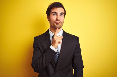 Takım elbiseli ve kravatlı genç yakışıklı bir işadamı izole edilmiş sarı arka planda dikilirken parmakları çenesinde olan şüpheyi düşünür ve yukarı bakar.