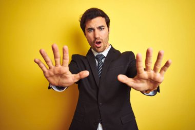 Takım elbiseli ve kravatlı genç yakışıklı işadamı izole edilmiş sarı arka planda durup avuç içi hareketlerini durduruyor, kızgın ve hayal kırıklığı ifadesi takınıyor.