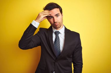 Takım elbiseli ve kravatlı genç yakışıklı işadamı, izole edilmiş sarı arka planda duruyor. Alnındaki sorun yüzünden endişeli ve stresli. Kriz konusunda endişeli.