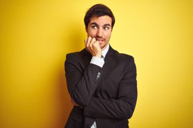 Takım elbiseli ve kravatlı genç yakışıklı işadamı, izole edilmiş sarı arka planda duruyor. Ağız ısıran elleri yüzünden gergin ve stresli görünüyor. Endişe sorunu.
