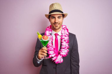İş adamı, takım elbiseli, Havai Lei marka bir şapka giyiyor. İzole edilmiş pembe arka planda kokteyl içiyor. Mutlu bir yüzü var ve gülümsüyor.