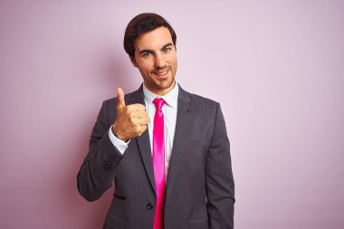 Takım elbiseli ve kravatlı genç yakışıklı bir işadamı pembe arka planda mutlu bir el hareketi yapıyor. Kameraya bakıp başarıyı gösteren ifade onaylanıyor.