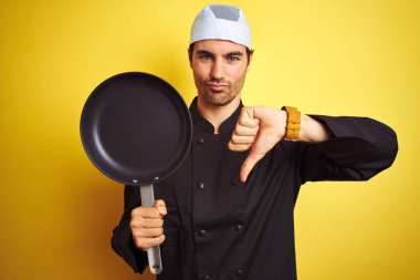 Üniformalı ve şapkalı genç aşçı tepsisi ile izole edilmiş sarı arka planda kızgın bir yüz, olumsuz işaret baş parmakları aşağıda, reddedilme konsepti var.