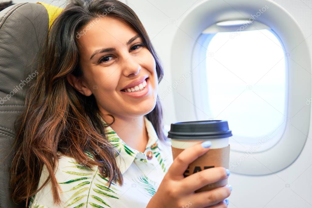 Young traveller woman sitting inside plane at the airport with sky view from the window drinking a cup of coffee