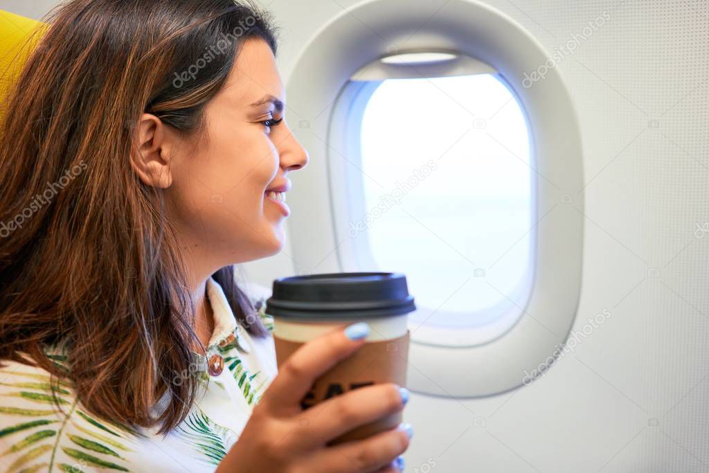 Young traveller woman sitting inside plane at the airport with sky view from the window drinking a cup of coffee