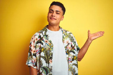 Tatilde olan genç Brezilyalı bir adam, izole edilmiş sarı arka planda çiçekli gömlek giyiyor, gülümsüyor ve avuç içi kameraya bakıyor..