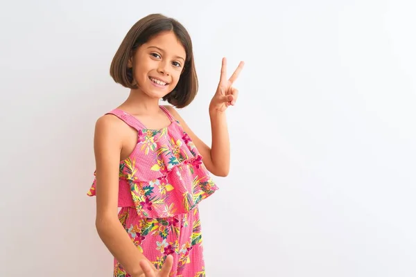 年轻美丽的小女孩穿着粉红色的花裙子 站在孤独的白色背景上 微笑着看着摄像机 手指头在做胜利的标志 第二点 — 图库照片