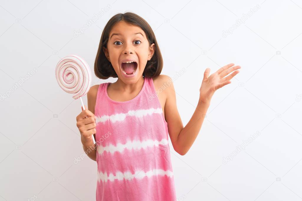Beautiful child girl eating sweet lollipop standing over isolated white background very happy and excited, winner expression celebrating victory screaming with big smile and raised hands