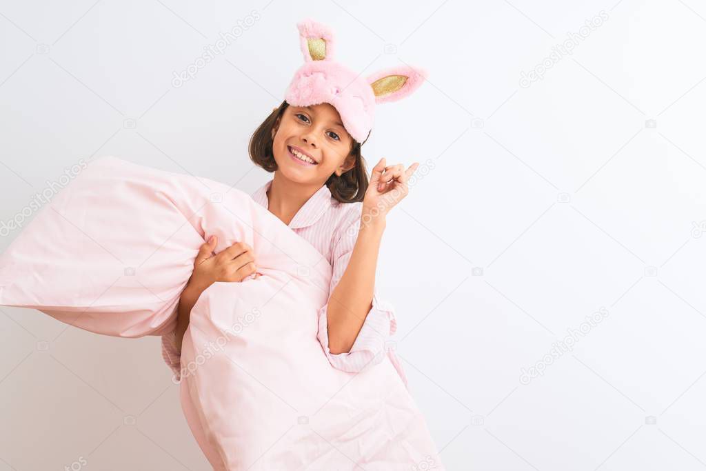 Child girl wearing sleep mask and pajama holding pillow over isolated white background surprised with an idea or question pointing finger with happy face, number one
