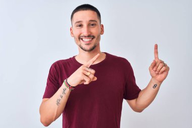 Genç yakışıklı adam izole edilmiş arka planda duruyor gülümsüyor ve kameraya bakıyor iki eliyle ve parmaklarıyla kenarı işaret ediyor..