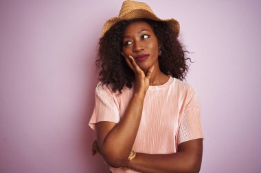 Genç Afro-Amerikalı kadın tişört ve şapka giyip pembe arka planda dolaşırken kollarını kavuşturup depresyondan sıkılmış ve yorgun görünüyor..