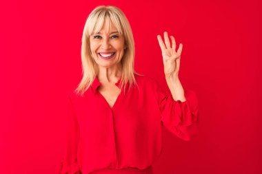 Soyutlanmış kırmızı arka plan üzerinde duran zarif gömlekli orta yaşlı bir kadın dört numaralı parmaklarıyla işaret ederken kendinden emin ve mutlu gülümsüyor..