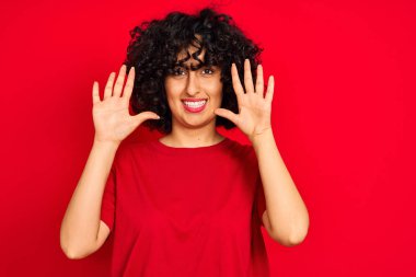 Kıvırcık saçlı genç Arap kadın izole edilmiş kırmızı arka plan üzerine günlük tişört giyiyor ve parmaklarını on numarayı gösterirken kendinden emin ve mutlu bir şekilde gülümsüyor..