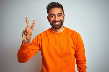 İzole edilmiş beyaz arka planda turuncu süveter giyen genç Hintli adam iki numaralı parmaklarıyla işaret ederken kendinden emin ve mutlu gülümsüyor..