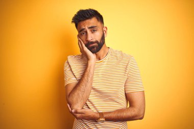 Sarı arka planda duran tişörtlü genç Hintli adam çapraz kollu depresyondan sıkılmış ve yorgun görünüyor..