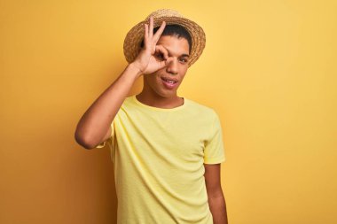 Genç yakışıklı Arap adam tişört ve yaz şapkası giyerek izole edilmiş sarı arka planda el hareketleriyle gülümsüyor ve mutlu yüzle parmaklarının arasından bakıyor..