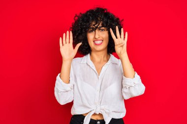 Kıvırcık saçlı, beyaz tişörtlü, izole edilmiş kırmızı arka planı olan genç bir Arap kadın dokuz numarayı gösterirken kendinden emin ve mutlu bir şekilde gülümsüyor..