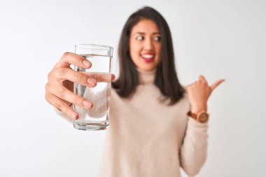 Genç, güzel bir Çinli kadın, elinde bir bardak su tutarak izole edilmiş beyaz arka planda duruyor ve gülümseyen bir gülümsemeyle baş parmağıyla onu gösteriyor.
